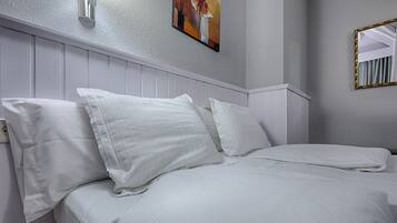 A neatly made bed with white linens and pillows in a room with white paneled walls and a painting above the bed.