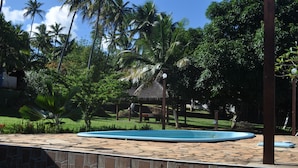 Una piscina con borde de piedra, rodeada de palmeras y una estructura con techo de paja.