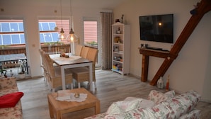 A living room with a sofa, a wooden coffee table, a dining table with chairs, a bookshelf, and a TV mounted on a wooden beam.