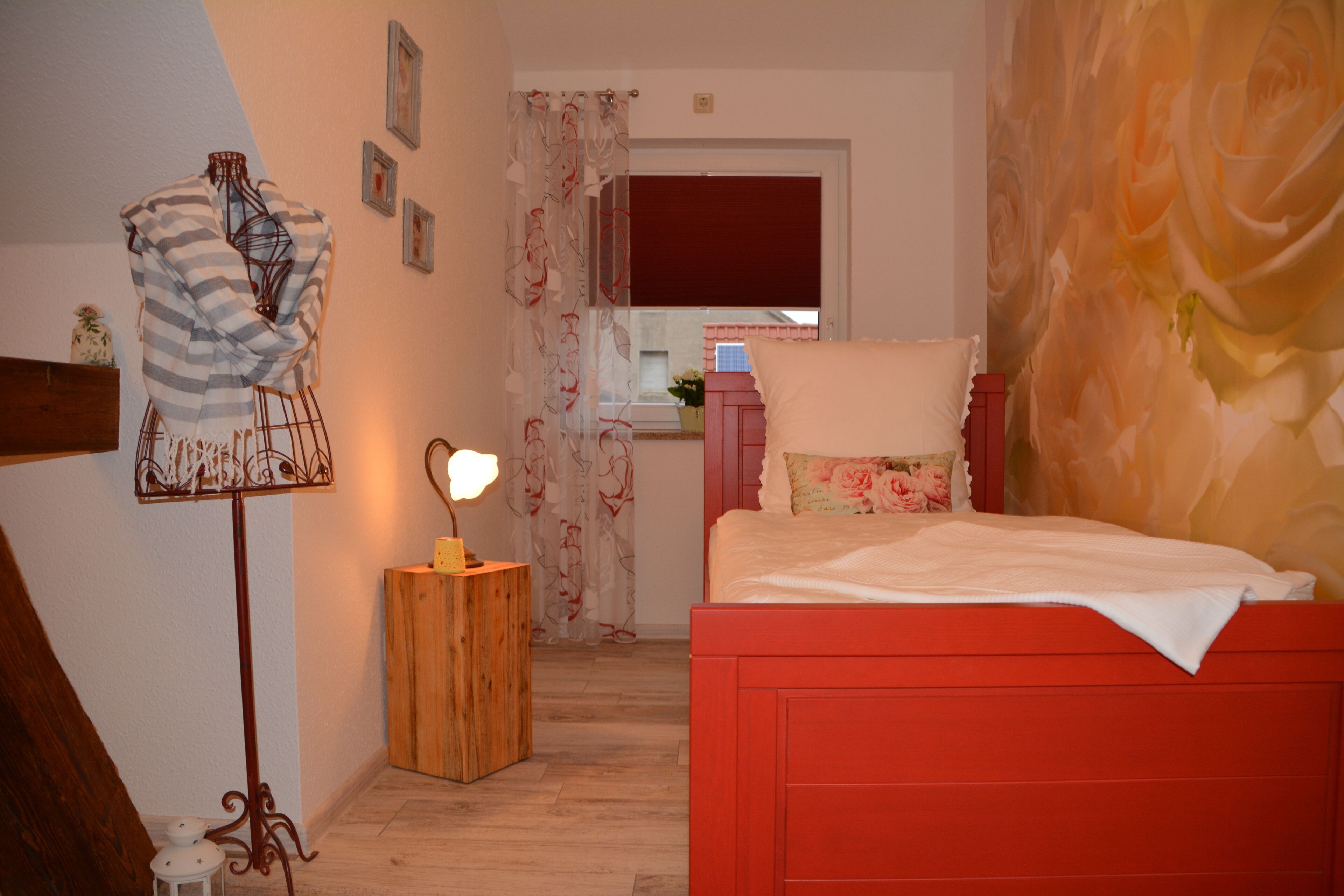 A bedroom with a red bed, a wooden nightstand with a lamp, a mannequin, and a window with a view of the outside.