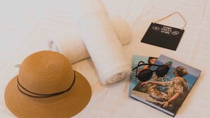 Ein Bett mit weißer Bettwäsche, ein Strohhut, ein gefaltetes Handtuch, Sonnenbrillen und ein Buch.