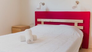 Ein ordentlich bezogenes Bett mit weißer Bettwäsche und einem aufgerollten Handtuch, ein Nachttisch aus Holz und ein leuchtend rotes Kopfteil.