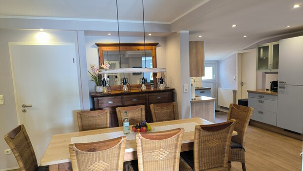 A dining area with a wooden table and wicker chairs, a kitchen with white cabinets, and a modern pendant light fixture.