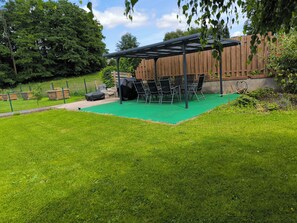 A covered patio area with a green artificial turf, surrounded by a wooden fence and lush greenery.
