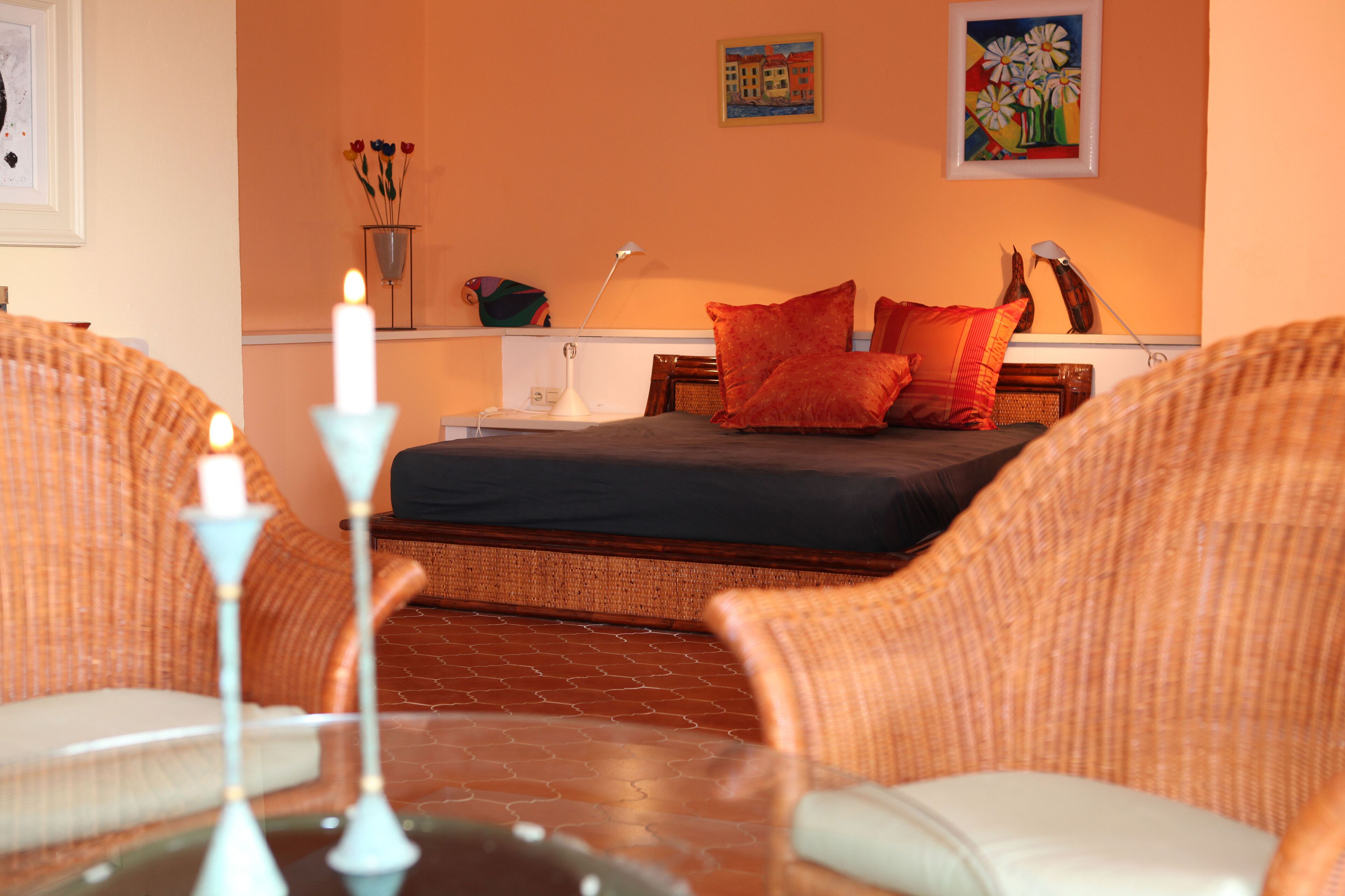 A room with wicker furniture, a bed with red pillows, a white candle, and artwork on the walls.