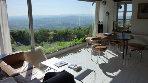 Une pièce spacieuse dotée d’une grande fenêtre offrant une vue panoramique sur la campagne. On y trouve une table à manger en bois avec des chaises, un canapé et une petite table sur laquelle sont posés des livres et une lampe.