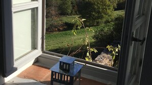 Une vue depuis une fenêtre, avec une petite table et une bougie, donnant sur un paysage verdoyant parsemé d’arbres et sur des collines lointaines.