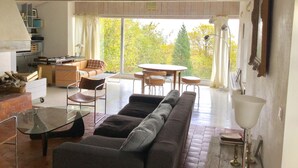 Un vaste espace de vie comprenant un grand canapé, une table basse en verre et une table à manger avec des chaises. Il y a également un coin cuisine avec une cuisinière et des placards, ainsi qu’une grande fenêtre donnant sur les arbres.