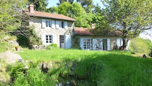 Une maison en pierre avec un toit de tuiles, un étang au premier plan et une végétation luxuriante tout autour.