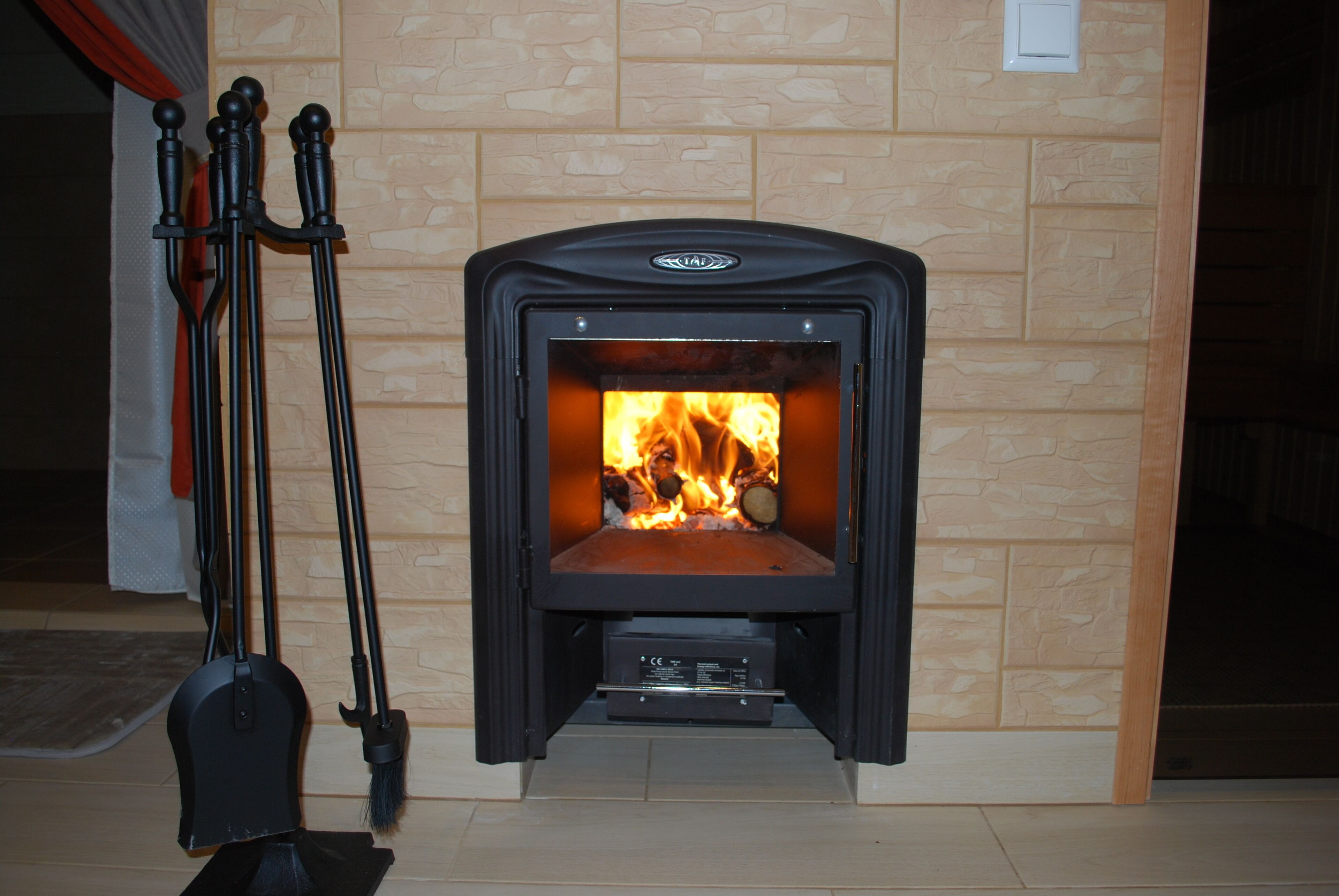 A wood-burning stove with a stone wall backdrop, a broom, and a shovel to the left.