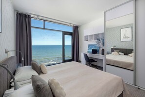 Ein Hotelzimmer mit Bett, Schreibtisch, Stuhl, einem großen Fenster mit Vorhängen und Blick aufs Meer.