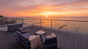 Blick auf einen Sonnenuntergang von einer Terrasse mit Außenbestuhlung und einem Geländer mit Meerblick.