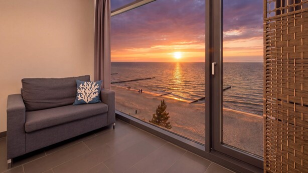 Ein modernes Zimmer mit großem Fenster und Blick auf einen Strand bei Sonnenuntergang, einem grauen Sofa und einem gewobenen Raumteiler.
