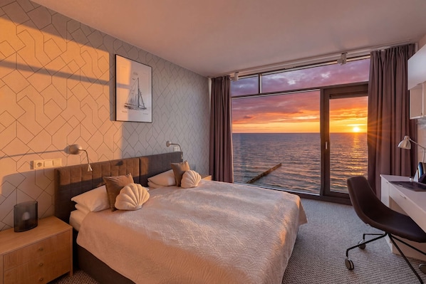 Ein Hotelzimmer mit Bett, Schreibtisch, Stuhl, Nachttisch, Lampe, einem Bild an der Wand und Blick auf den Sonnenuntergang über dem Wasser.