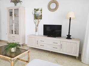 Ein Wohnzimmer mit Fernseher, Schrank, Lampe und einem Glastisch mit Pflanze.