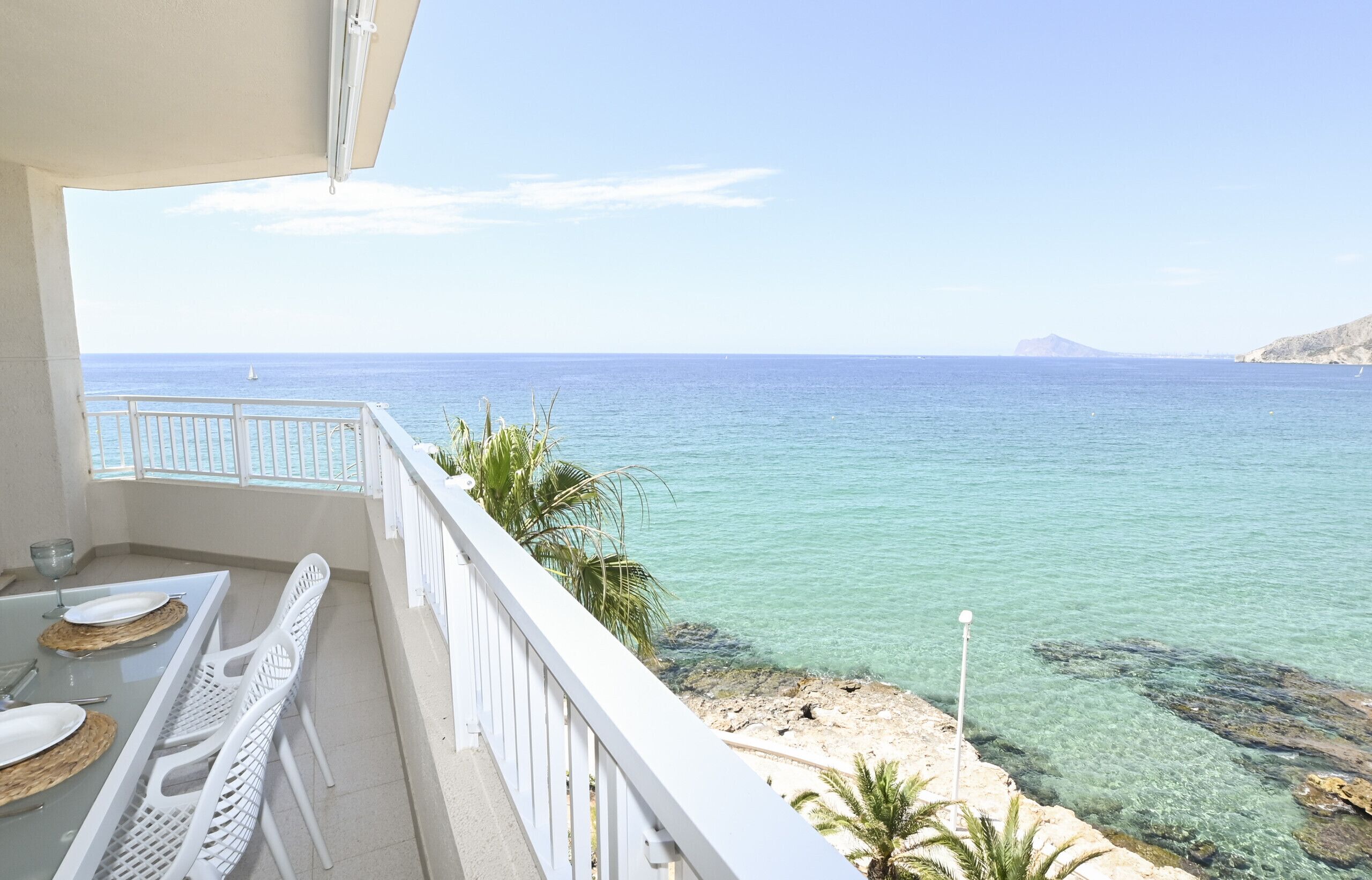 A balcony with a clear view of the sea, a dining table with chairs, and a white railing.