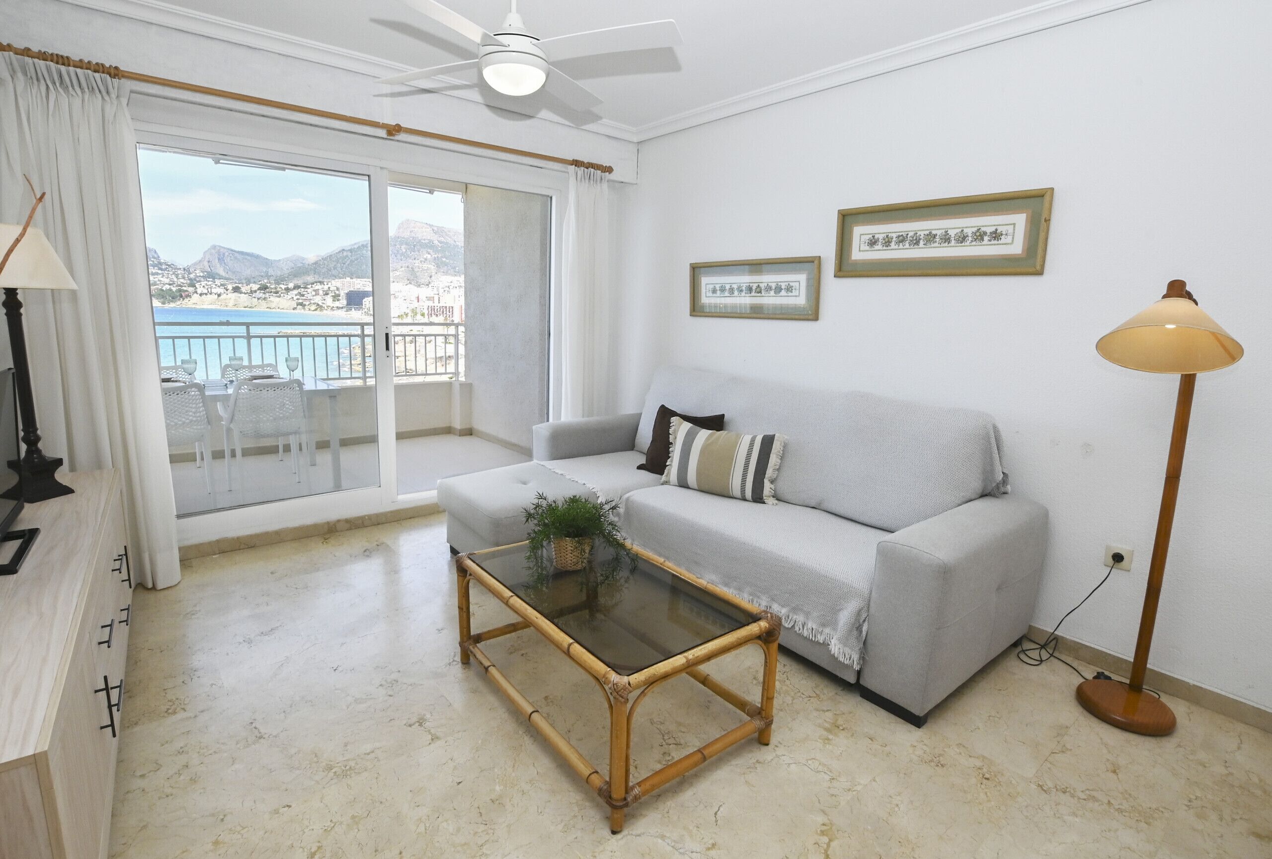 A living room with a glass-top coffee table, a grey sofa, a ceiling fan, and a view of the sea through a balcony.