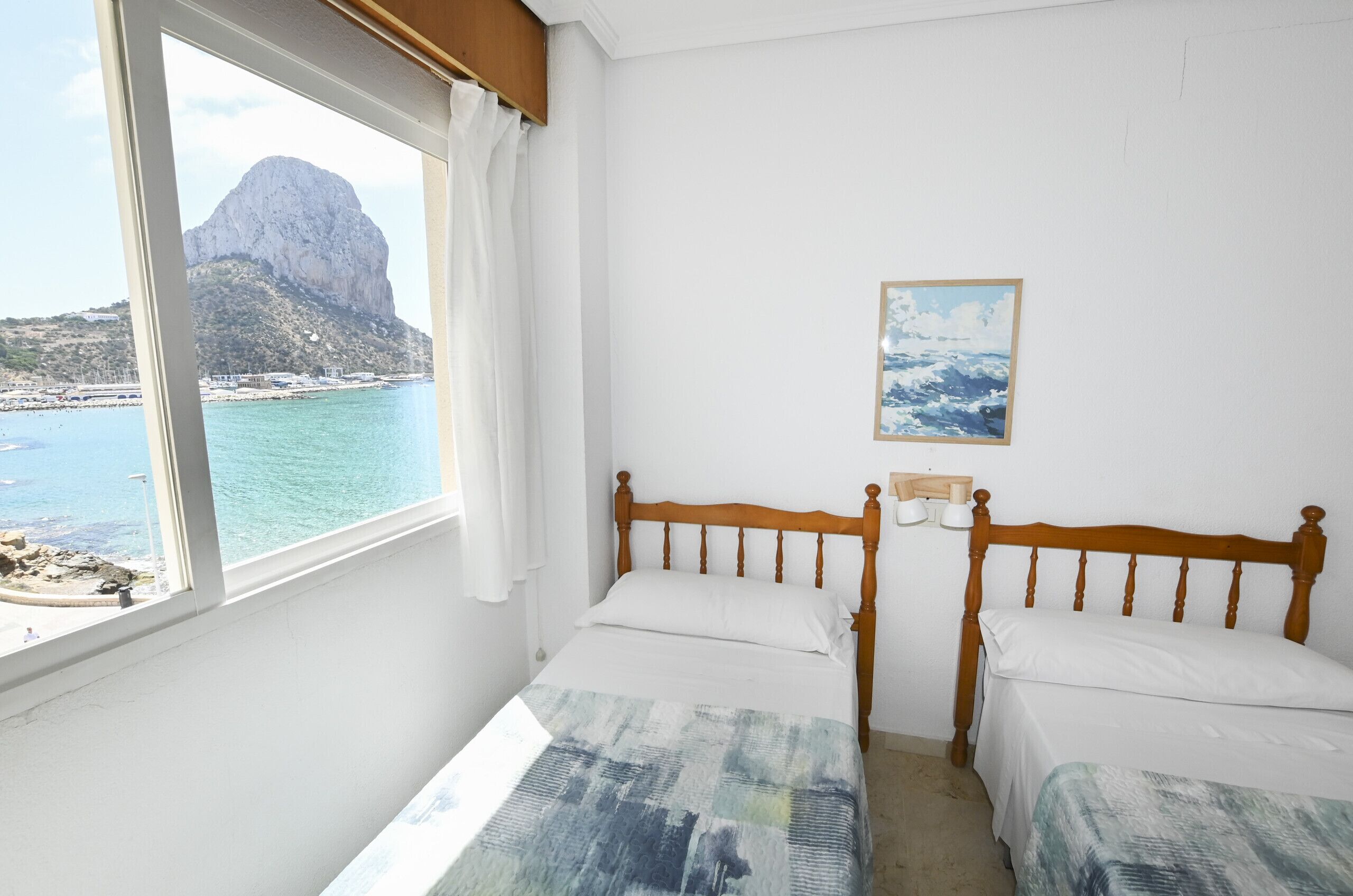 Une chambre avec un lit, une fenêtre donnant sur une vue de montagne et de mer, et un tableau accroché au mur.