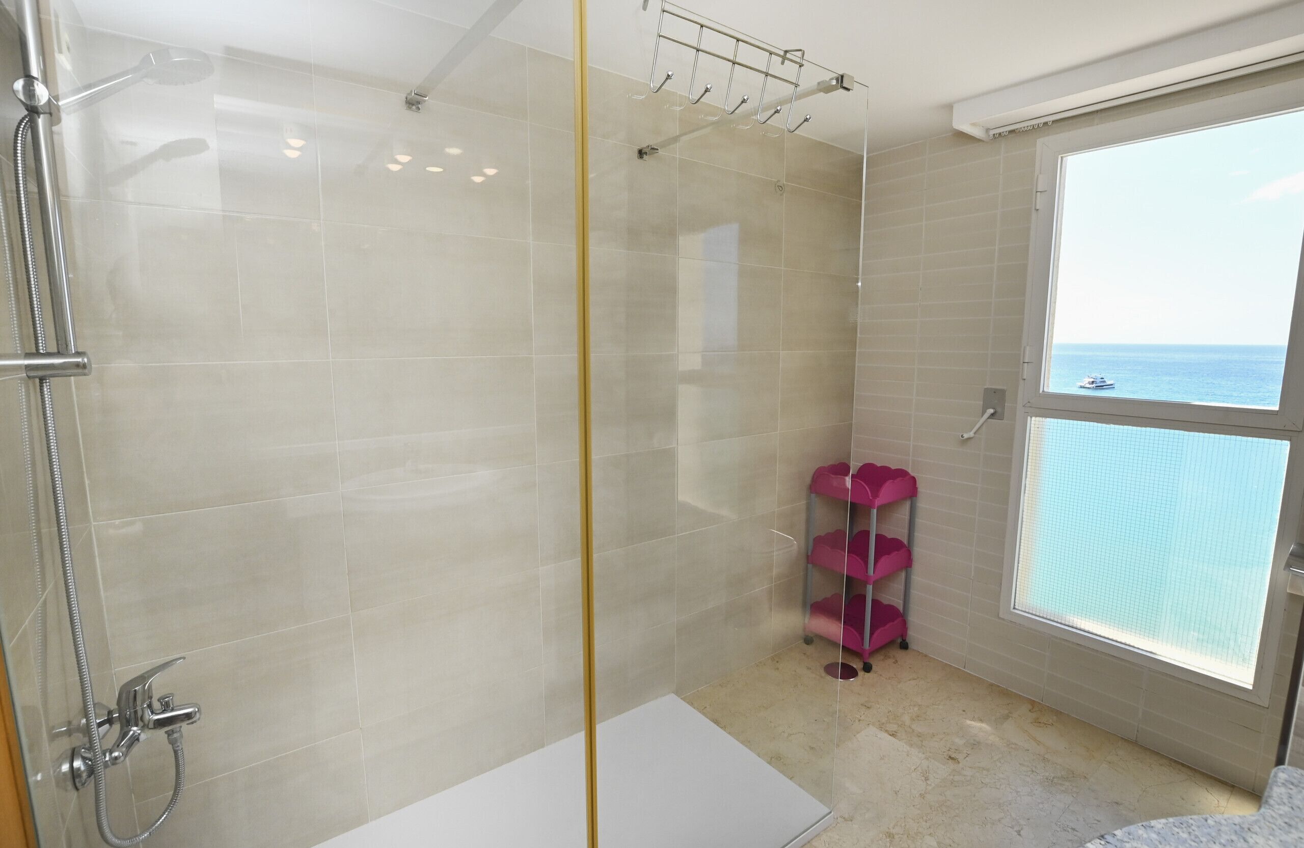 A modern bathroom with a glass shower enclosure, a window with a view of the sea, and a pink storage unit.