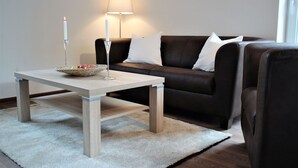 Ein Wohnzimmer mit einem Holztisch, einer Lampe, einem Obstkorb und einem dunkelbraunen Sofa mit weißen Kissen.