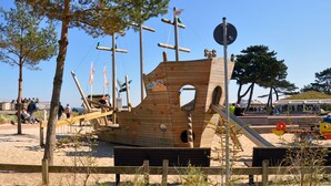Una estructura de barco pirata de madera con puntos de escalada y un tobogán, rodeada de árboles y una playa de arena.