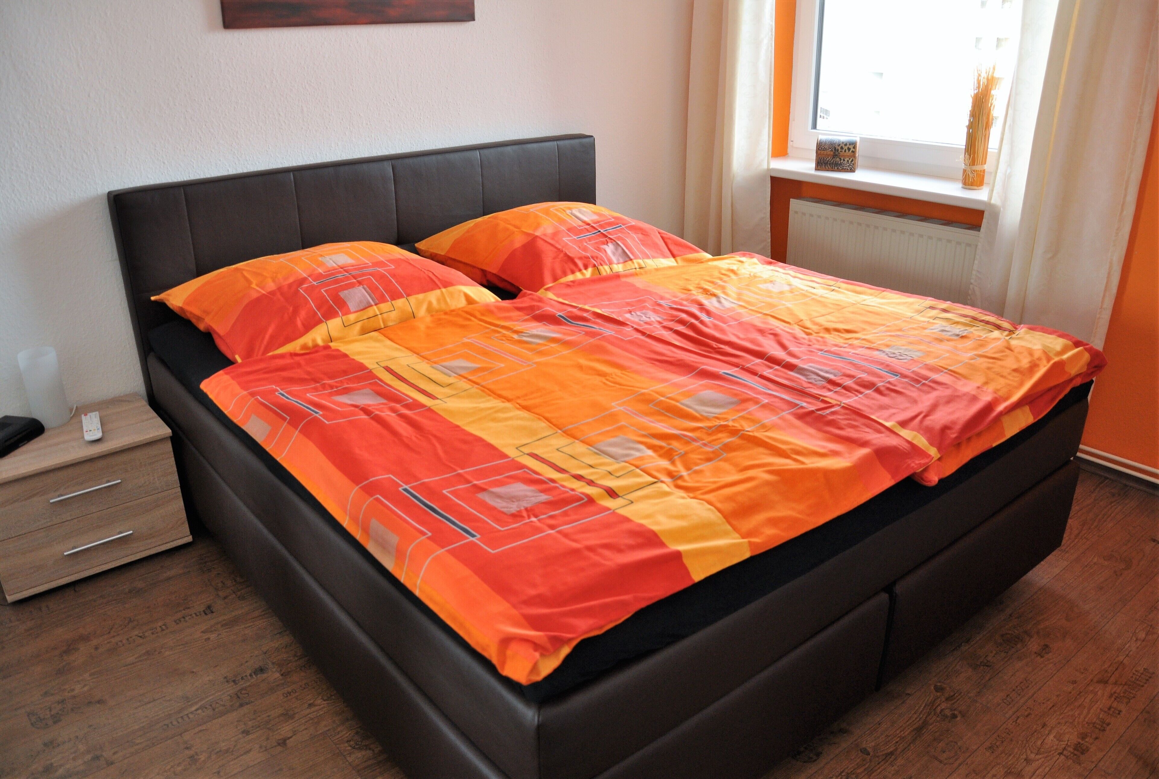 Una cama con cabecera negra y ropa de cama naranja, una mesita de noche de madera con cajones, y una ventana con vista a la calle.