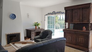 Ein Wohnzimmer mit Kamin, einer Couch, einem Sessel, einem Schrank und Blick ins Freie.