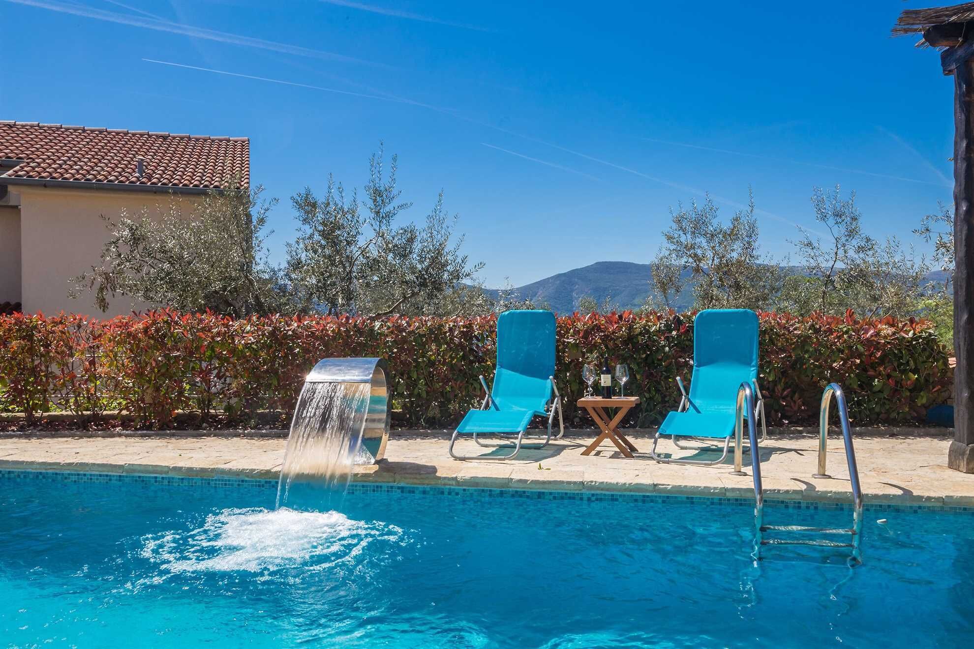 A swimming pool with a waterfall feature, two blue lounge chairs, and a table with wine glasses.
