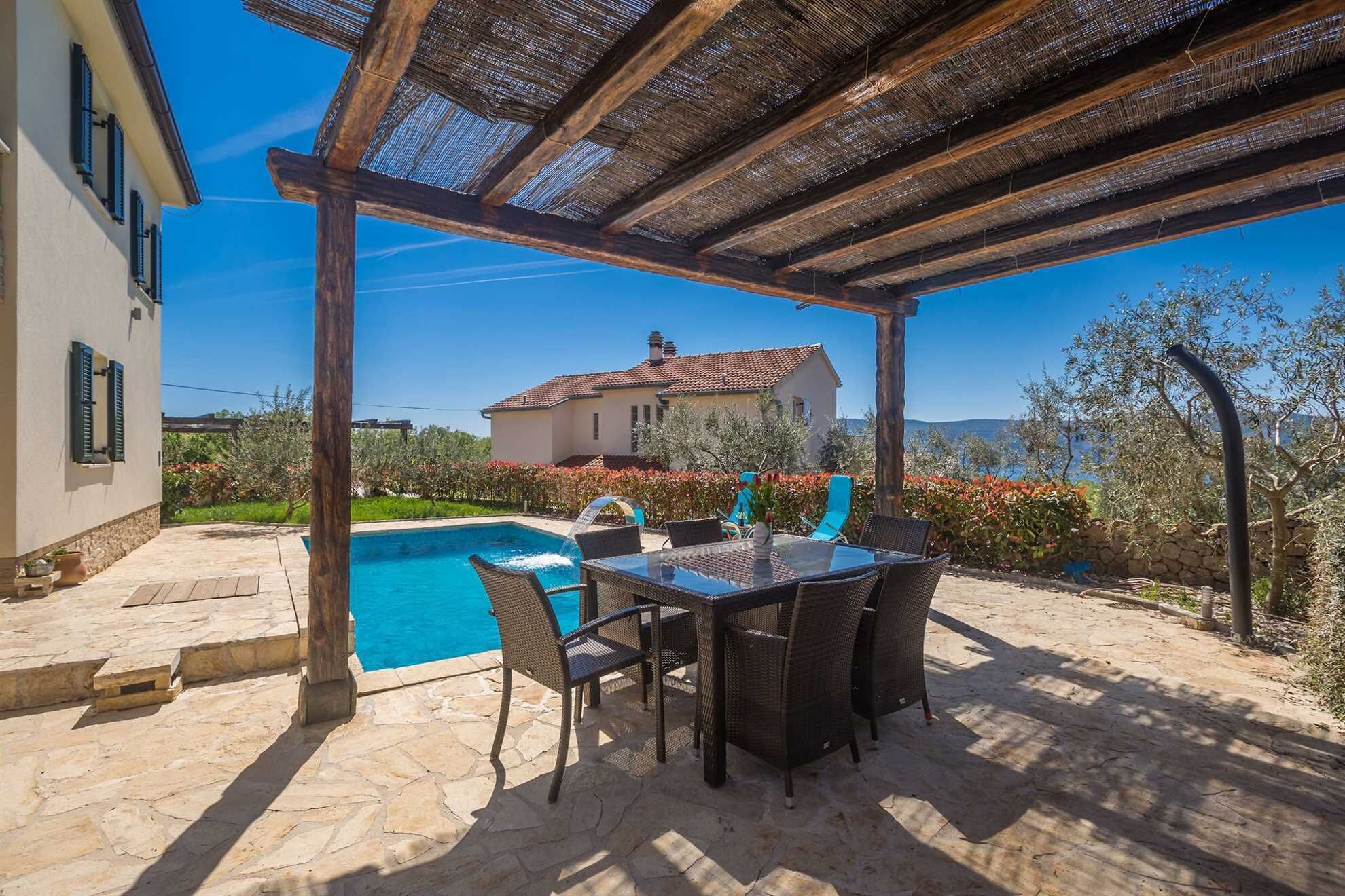 A covered outdoor area with a pool, wicker furniture, and a view of a house in the background.