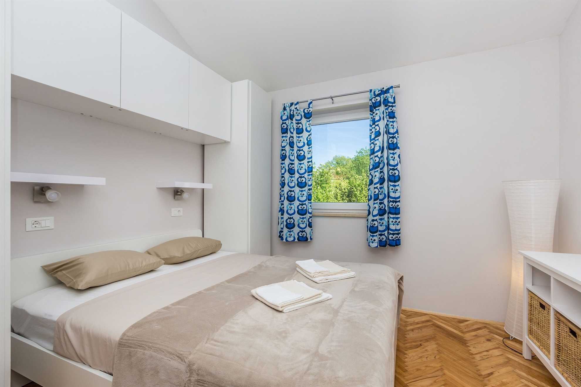 Ein Schlafzimmer mit einem Bett, zwei Kissen, einem Fenster mit blauen Vorhängen, einem Nachttisch und einer Lampe.