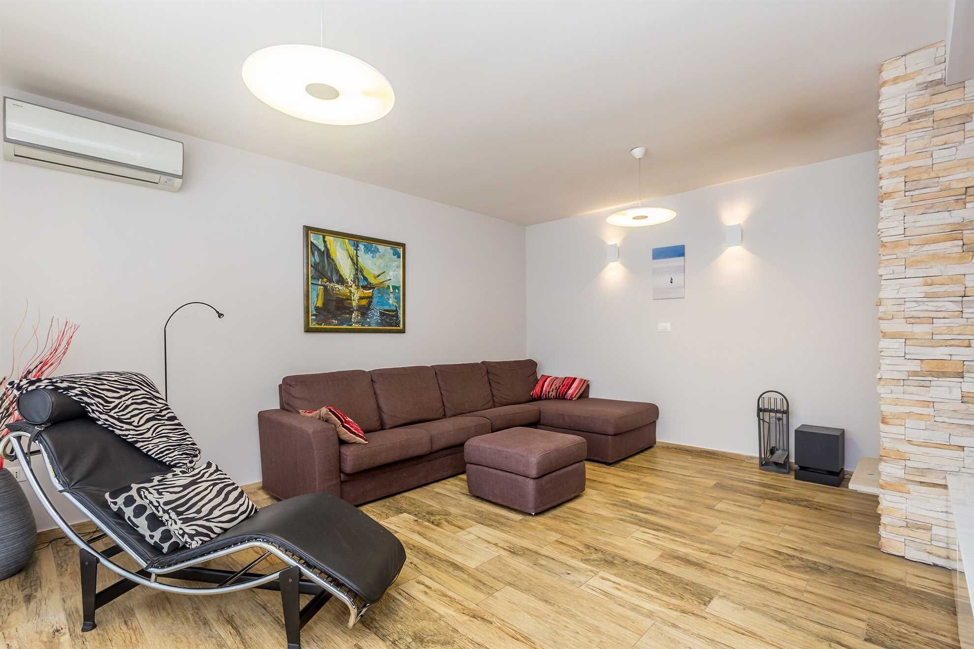 Un salon moderne avec un canapé d’angle, une chaise longue et une petite table. On y trouve un tableau au mur, une cheminée et un mur d’accent en pierre.