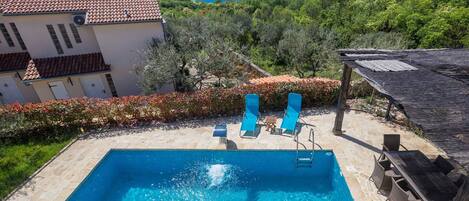 En swimmingpool med krystalklart, blåt vand, omgivet af stentile. Liggestole og en overdækket træpergola. Poolen ligger ved siden af en bygning med et tag af fliser og hvide vægge.