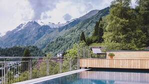 A swimming pool with a wooden deck and metal railing, surrounded by lush greenery and mountains in the background.