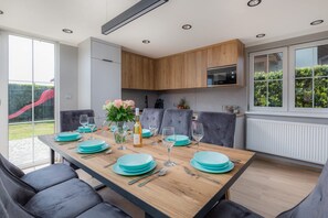 Un comedor moderno con una mesa de madera dispuesta para una comida, sillas grises tapizadas y una cocina con armarios de madera y un microondas integrado.