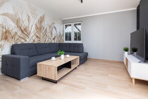 Ein modernes Wohnzimmer mit einem dunkelgrauen Ecksofa, einem Holztisch, einem Flachbildfernseher auf einem weißen Standfuß und einem großen Fenster mit Blick auf Grünflächen.
