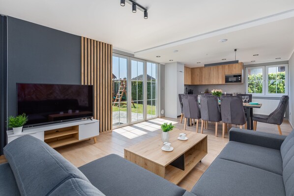 A modern living room with a flat-screen TV, grey sofas, a wooden coffee table, and a dining area with grey chairs.