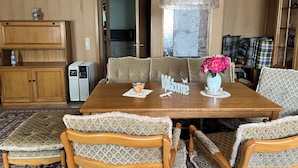 Una habitación con paneles de madera, con una mesa de comedor lista para cuatro personas, rodeada de sillas estampadas. Se ve una ventana grande con una parte superior de vidrio decorativa, y un jarrón con flores está sobre la mesa.