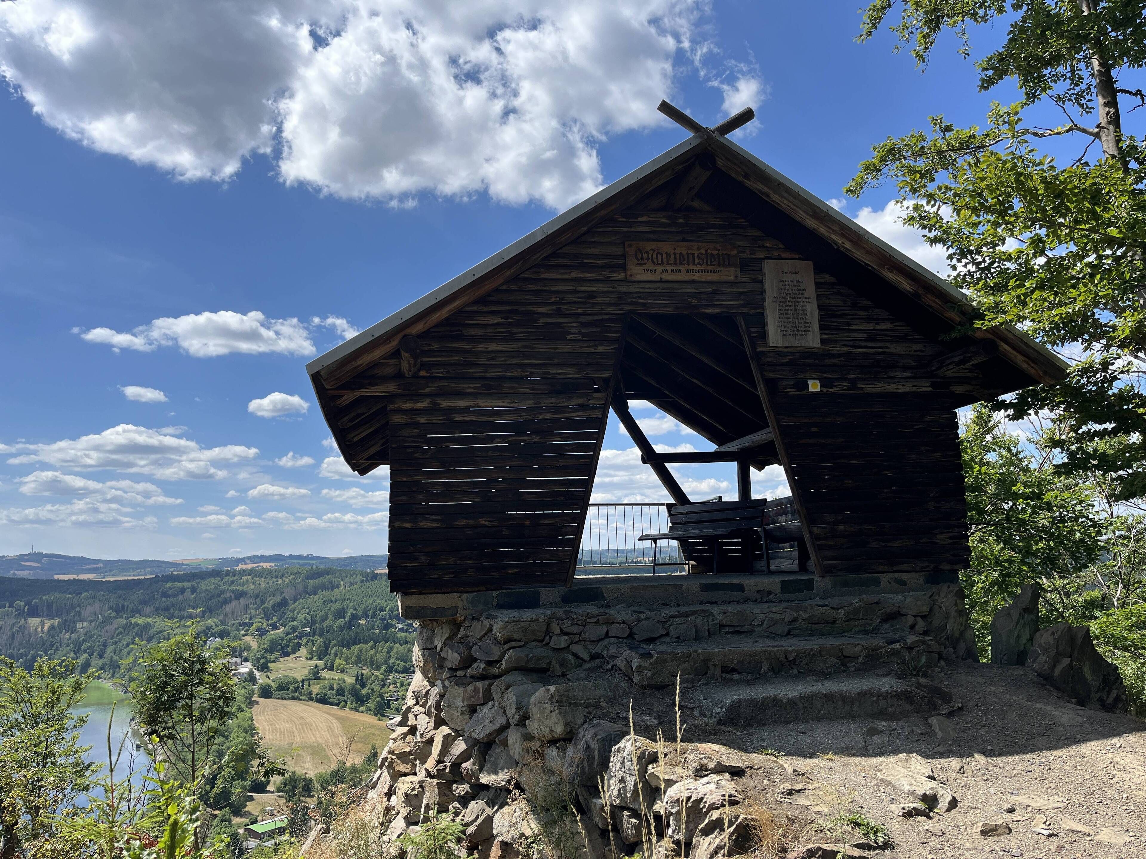 A wooden cabin with a stone base, a sign indicating its historical significance, and a view of the surrounding landscape.