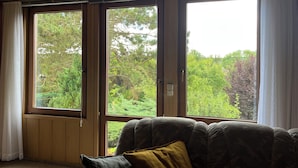 Una habitación con un gran ventanal corredera que da a un jardín, una zona de estar con cojines y techo de madera.