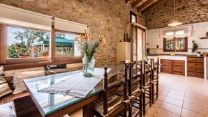 Un'area pranzo rustica con pareti in pietra, un tavolo in vetro e sedie in legno, una cucina con pensili in legno e vista all'esterno attraverso ampie finestre.