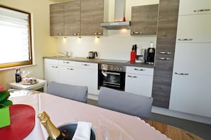 Une cuisine moderne dotée d’appareils électroménagers intégrés, un coin repas avec une table dressée pour deux personnes, et une fenêtre équipée de stores.