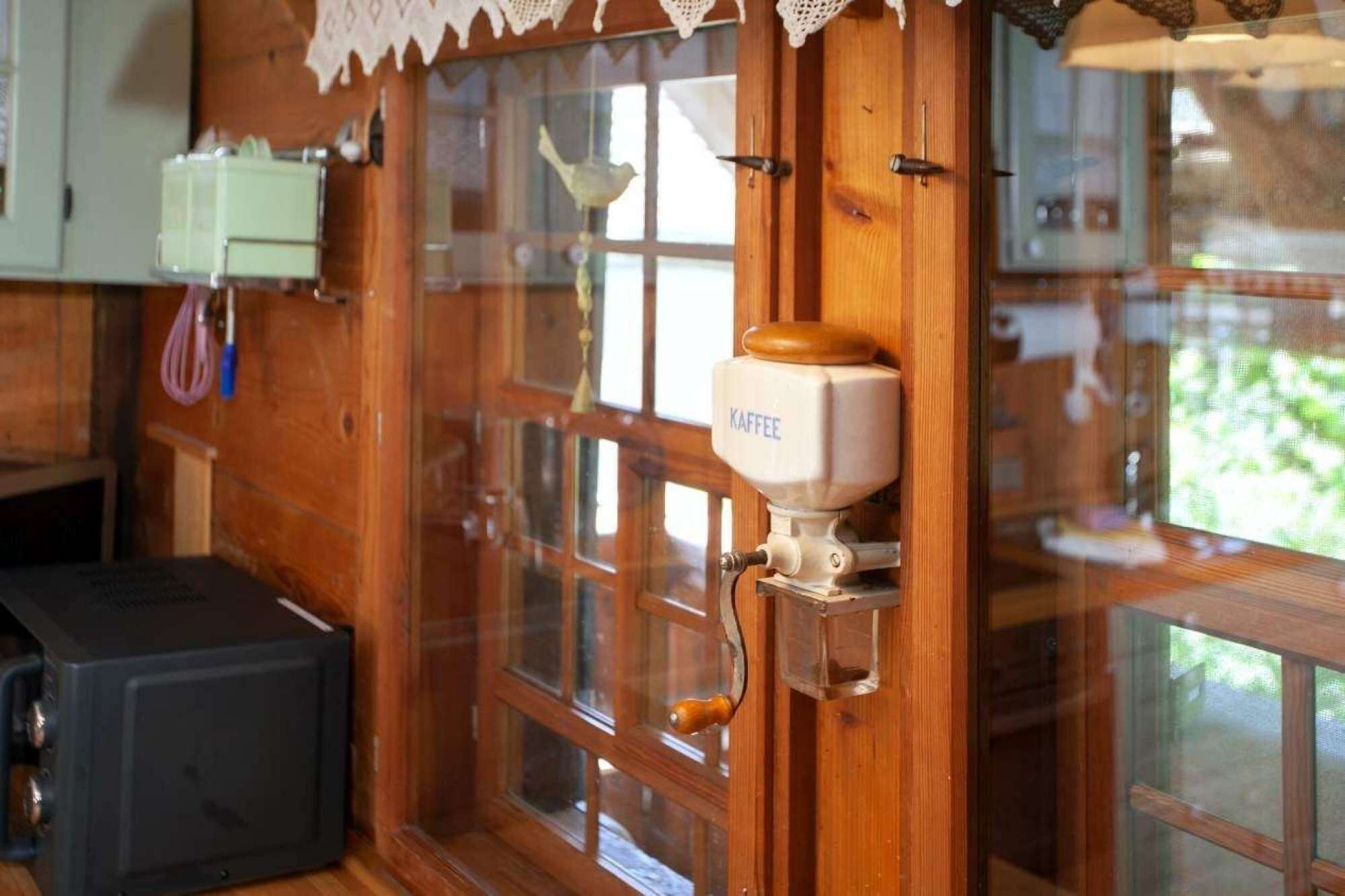A wooden interior with a coffee machine labeled "KAFFEE" mounted on a wall, a window with a view of greenery, and a small cabinet with a green appliance.