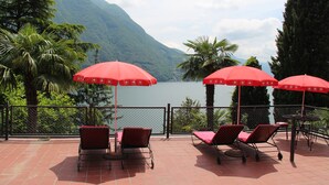 Una terraza con sombrillas rojas y sillones de descanso con vista a un lago y montañas al fondo.