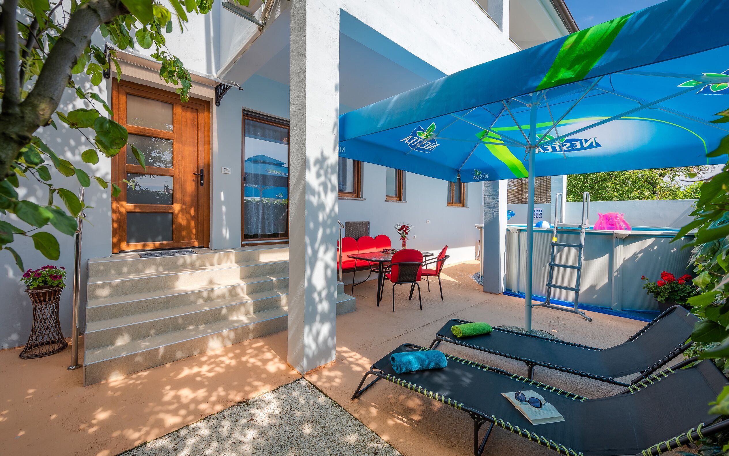 Eine sonnendurchflutete Terrassenanlage mit Pool, roter Sitzgelegenheit und einer Lounge-Bereich mit blauem Sonnenschirm.