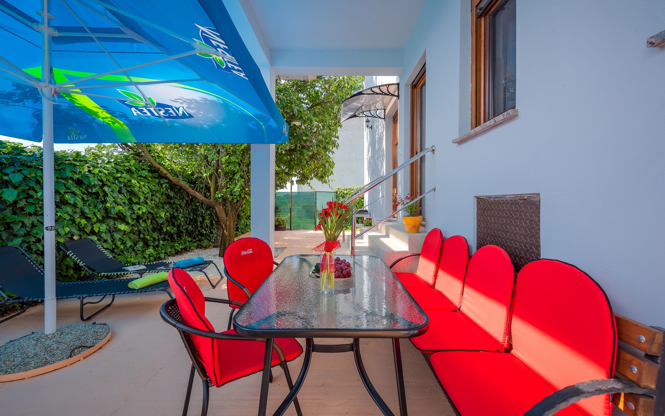 An outdoor seating area with a glass table and red chairs, a blue umbrella, and a hedge in the background.
