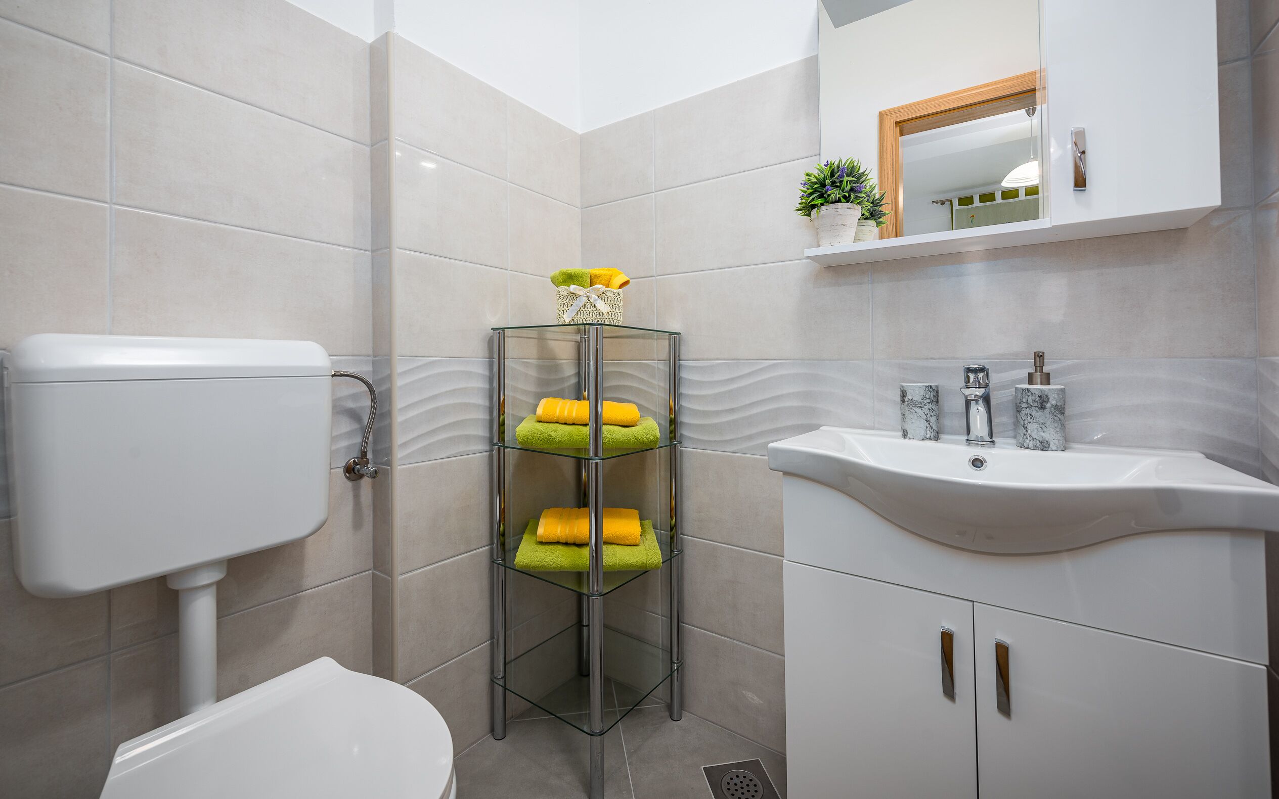 Une salle de bain moderne comprenant des toilettes, une étagère en verre avec des serviettes, un lavabo avec un socle en marbre et un miroir au-dessus du lavabo.