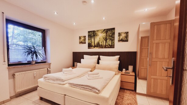 Ein modernes Schlafzimmer mit einem großen Bett, einem Nachttisch, einem Fenster mit Blick auf Bäume und einer Tür.