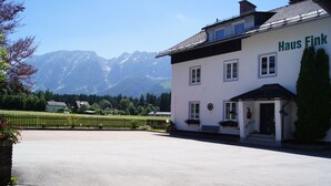 A white building named "Haus Fink" with a balcony, a paved driveway, and a mountain backdrop. - Apartment avalanche stone (Bad Mitterndorf)