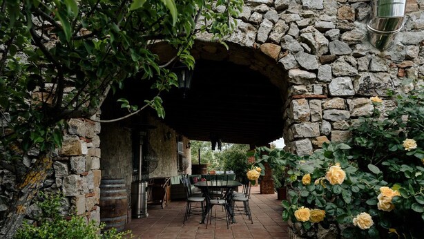 A stone archway leading to a patio with a table and chairs, surrounded by greenery and yellow roses.