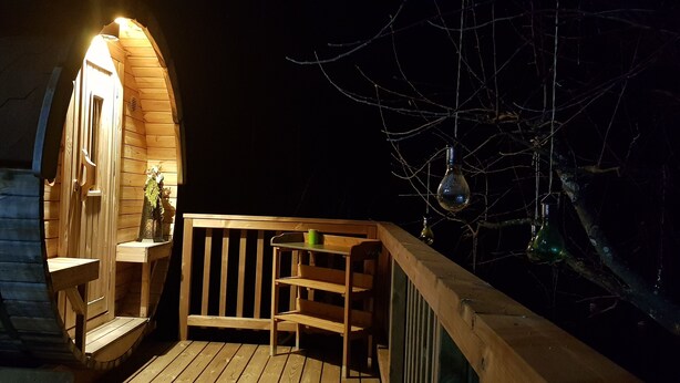 En træterrasse med bænk og lille bord, en trækonstruktion med skråt tag samt et træ med hængende pynt.